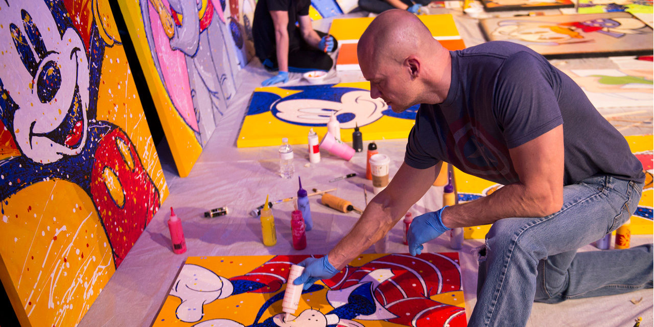Man on one knee leaning over to use squeeze-bottle of white paint on a Disney character painting for Epcot International Festival of the Arts.