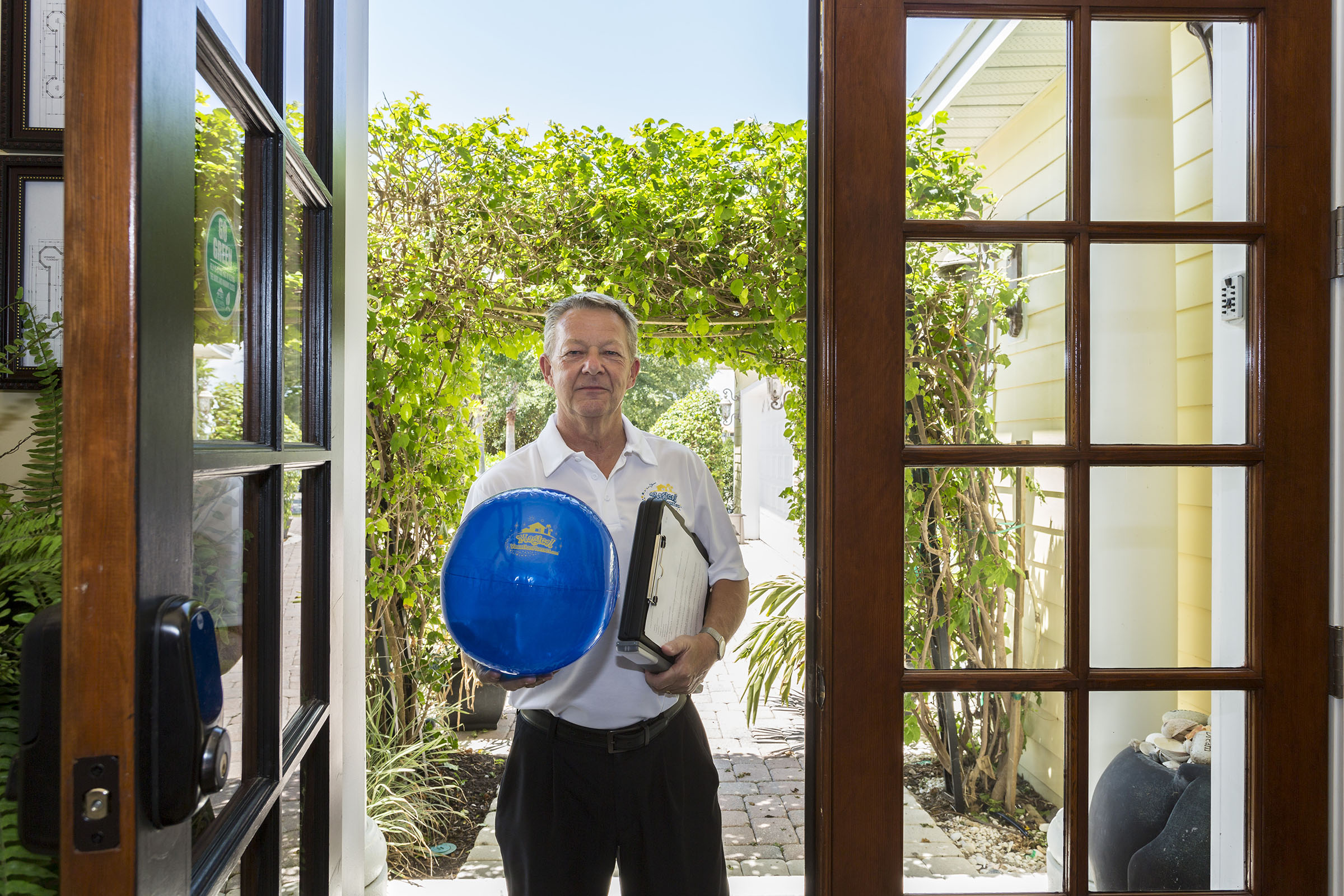 Older man standing at outside entrance of brown french doors holding a see-through royal blue beach ball with yellow writing of logo "Magical Vacation Homes". Archway made of vines. Daytime photo.