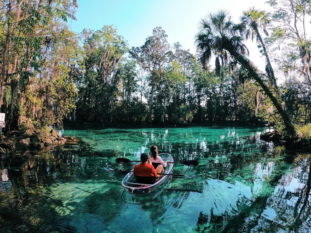 A man in back of kayak and a woman in front of kayak (clear kayak) floating in a natural turquoise spring through land conservation.